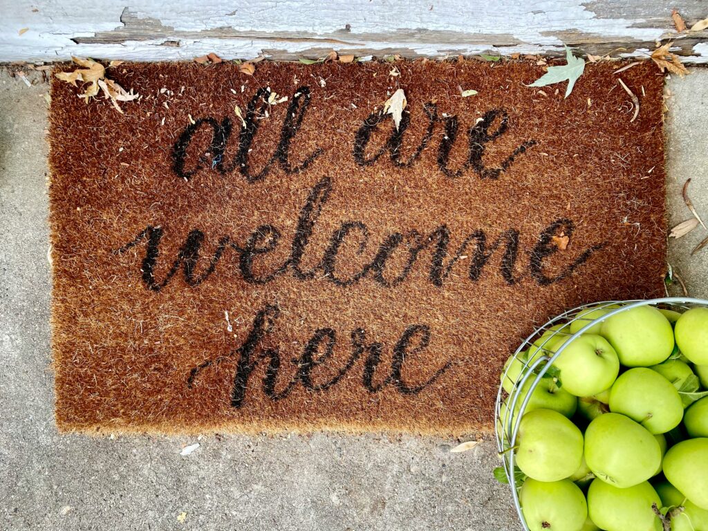 a welcome mat which reads “all our welcome here” with a metal container of green apples beside it.