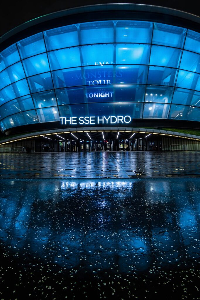 The Hydro at night. It is a large, round glass building lit with blue lighting. A sign outside reads “The SSE Hydro.” The ground outside is wet from rain.