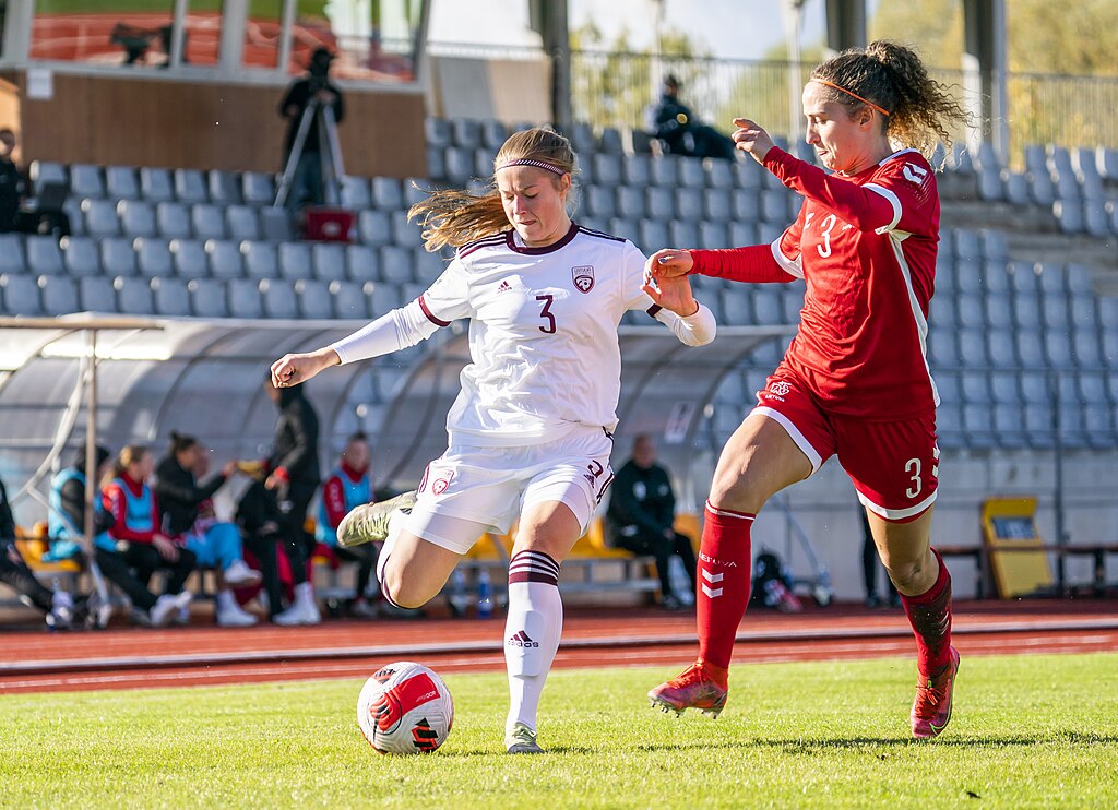 Paula Linda Liniņa representing Latvia national women's football team against Lithuania. One woman with light skin tone and long blonde hair is wearing a white football kit and is about to kick a football, right beside her is a woman with light skin tone and wavy brown hair wearing a red football kit who is also going for the ball.