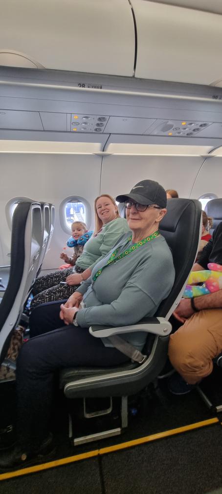 an aeroplane row with a small child sat by the window with a neck pillow round their neck and a lollipop in their hand, a woman with light skin tone and long blonde hair in the middle seat smiling, and a woman with light skin tone wearing a black baseball cap with a sunflower lanyard on in the aisle seat.