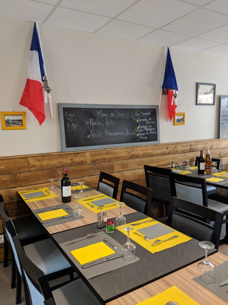 a restaurant in France, the tables are laid ready for people to arrive. On the wall is a chalkboard menu with a french flag either side showing “menu du jour; entre, buffet, moules frites, poulet Basquaise frites. Nos desserts; compote de pommes, salade de kiwi/pasteque, soupe de fruits rouge.”