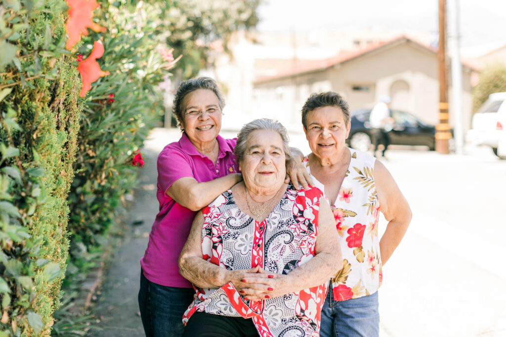 3 women of different ages with medium light skin tone and short hair standing outside beside a hedge on a bright sunny day, all smiling broadly at the camera.