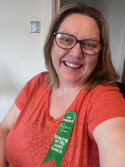 a woman with light skin tone and shoulder length light brown hair wearing glasses and smiling broadly at the camera. She is wearing an orange top with a green ribbon pinned to it which reads “got dressed rather than staying in PJs until lunch.”