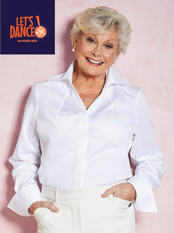 Angela Rippon, a older woman with light skin tone and short blonde hair, wearing a white shirt and white fitted trousers with her hands in the pockets. She is smiling at the camera. The Let’s Dance logo is in the top left corner.