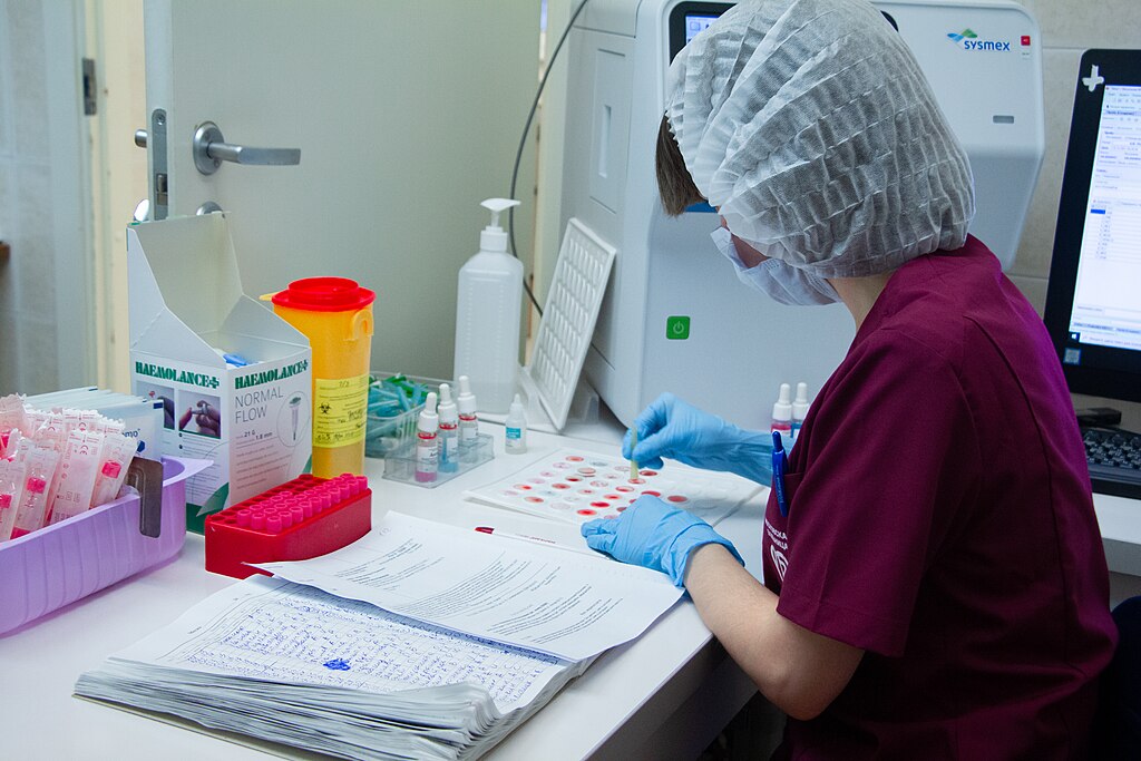 a person wearing a medical hair net, gloves and mask sitting at a table with lots of blood testing equipment on it and paperwork.