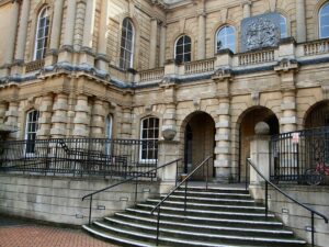 stairs lead up to Reading crown court, a stone building with archways at the front and tall windows on both the ground and first floors. A coat of arms is displayed on the outside of the first floor behind the balcony. Black railings run around the outside.