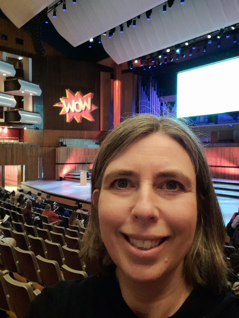 Sarah, a woman with light skin tone and shoulder length brown hair. Behind her is a large stage and many theatre chairs slowly filling up with people. On the wall above is the WOW of the Women of the World Foundation logo.