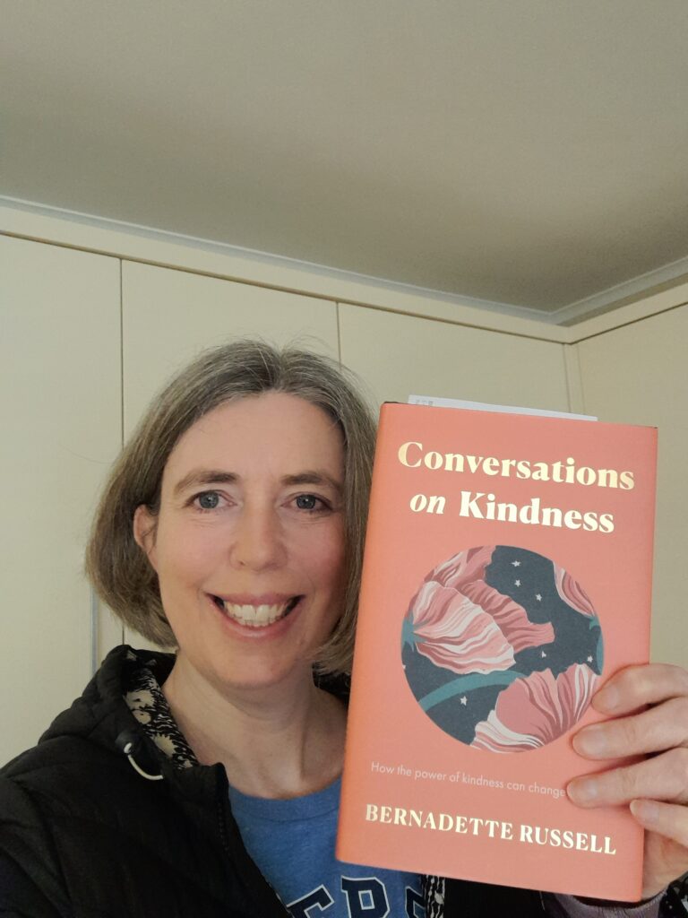 Sarah, a woman with light skin tone and short grey hair in a bob smiles broadly while holding a book with an orange cover and flowers in a central circle. The book is called “conversations on kindness” by Bernadette Russell