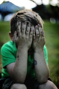 a young boy wearing a green tshirt, sits on a grassy floor, head in his hands, mud and scrapes down his arms.