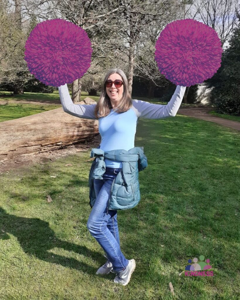 Sarah, a woman with light skin tone and shoulder length light brown hair, wearing sunglasses, jeans and a blue top with a jacket tied around her waist. She is standing in a park holding her hands in the air and two pink and purple pom poms have been overlaid onto them.