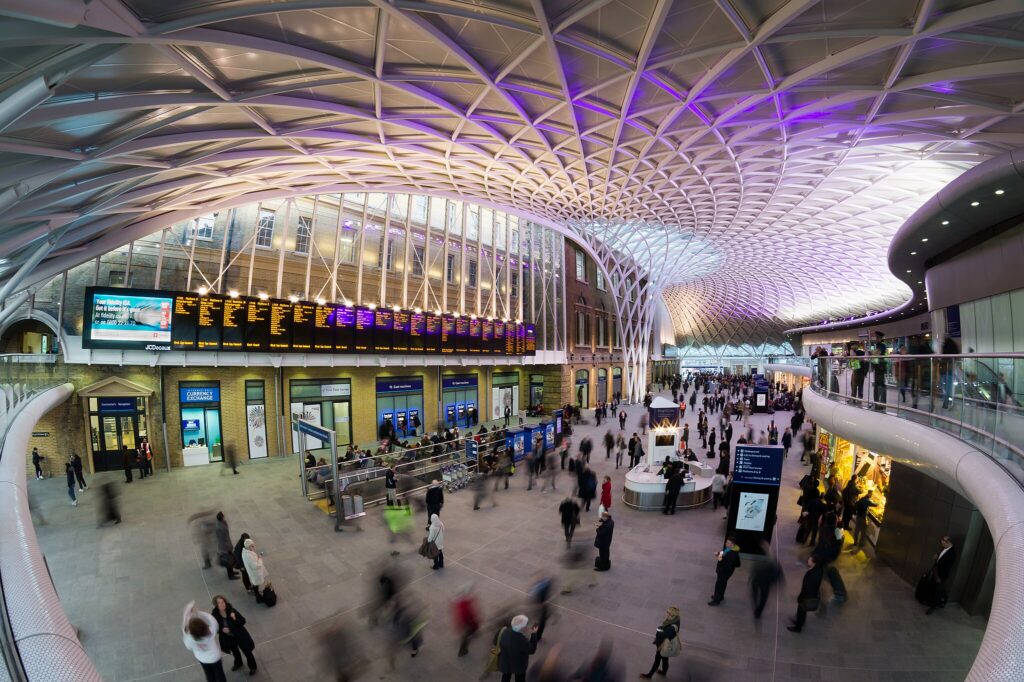 Kings Cross station with its artsy ceiling and lots of screens showing arrivals an departures on different platforms. At the left of the picture, below the walkway, is Platform 9¾ where a Harry Potter fan is posing for his picture. Most passengers are in a hurry to catch their train home; those dallying are engrossed in their mobile phones.