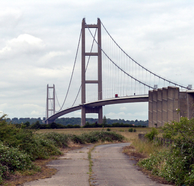 The Humber Bridge, a large suspension bridge, viewed from a countryside road beside one end of it. The sky above is cloudy and a few large vehicles are just visible on the bridge.