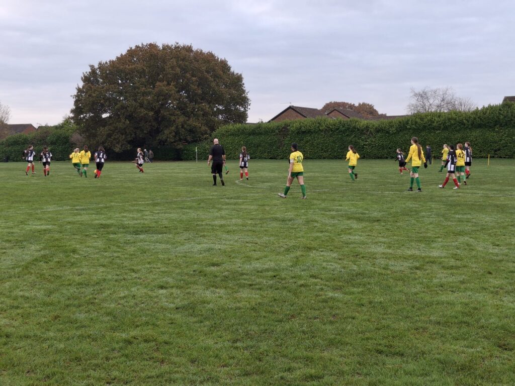 a grass football pitch with two women’s youth teams playing football on it. The sky is overcast.