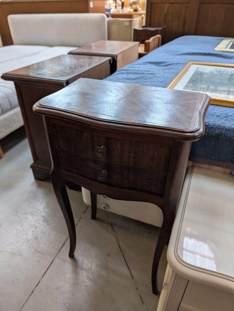 a wooden bedside table with slim shapely legs and two small draws with little round knobs. It is quite simple but looks beautiful and well made. Behind it are more items in the brocante.
