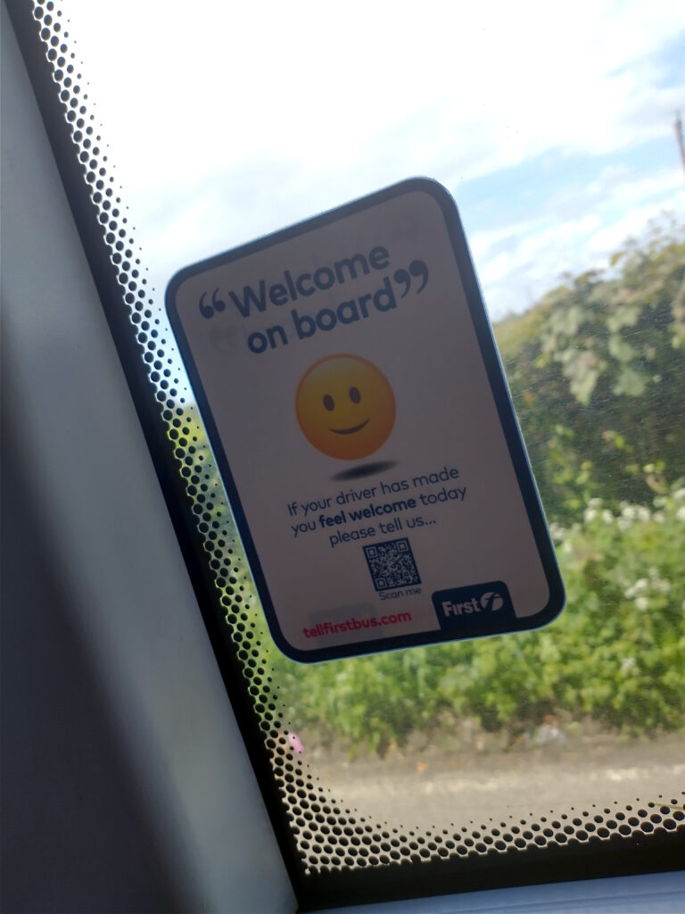 a sticker on the window of a bus which reads “welcome on board. If your driver has made you feel welcome today, please tell us.” There is a smiley face and a QR code also on the sticker.