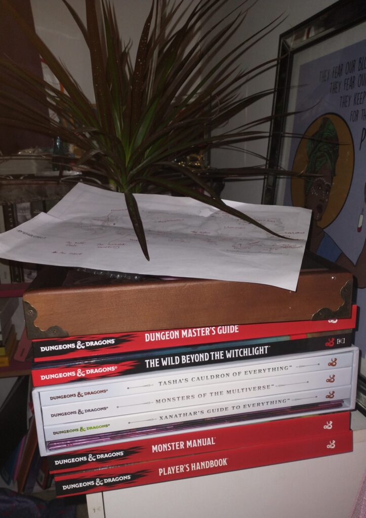 a pile of Dungeons and Dragons books on a table; player’s handbook, monster manual, Xanathar’s guide to everything, Monsters of the multiverse, Tasha’s cauldron of everything, The wild beyond the witchlight, and the Dungeon master’s guide. On top of them is a wooden dice rolling trey and on top of that a piece of paper. Behind it all is a plant.