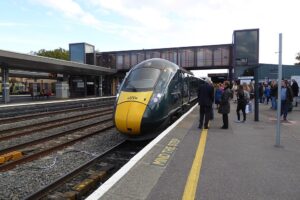 a train waiting at Oxford station. On the platform are many people waiting to board. The sky above is blue but with lots of white clouds.