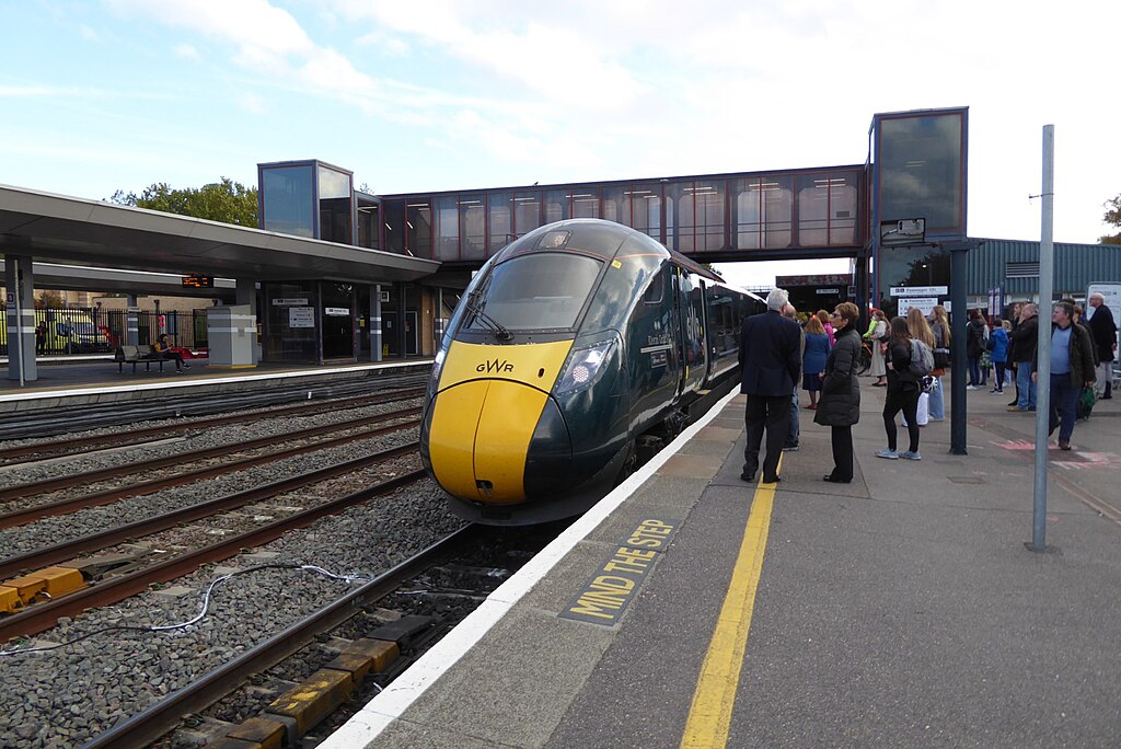 a train waiting at Oxford station. On the platform are many people waiting to board. The sky above is blue but with lots of white clouds.