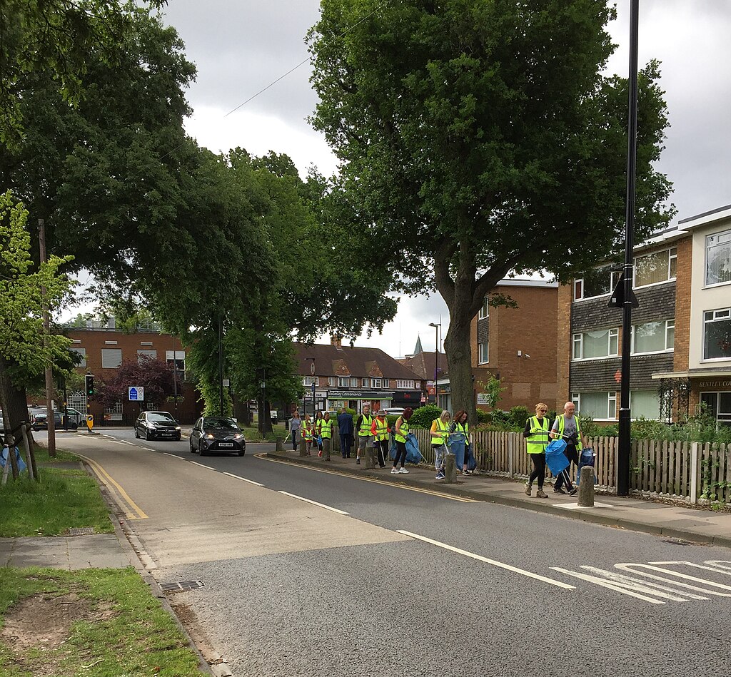 a group of litter pickers wearing high vis jackets and carrying bags and long handled picking devices walk along a pavement beside a road. There are some trees along side the route and buildings alongside.