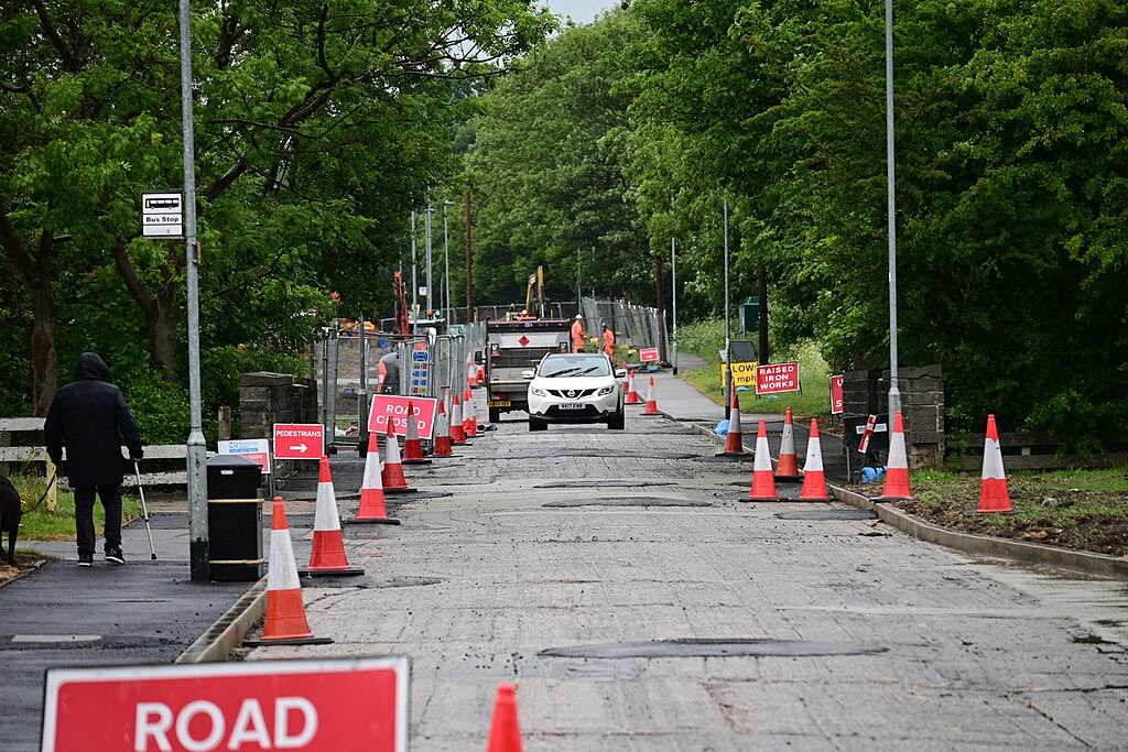 a road with cones on both sides of it. In the foreground is a road closed sign. On the road is a white car driving towards the camera which has just passed a flat bed truck. To the left someone is walking a dog along the pavement. There are trees either side of the road.