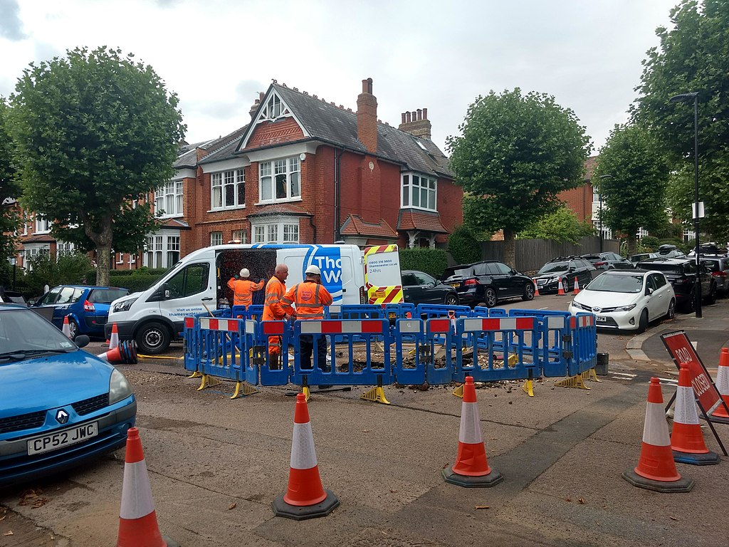 a cordoned off part of a residential road with traffic cones with blue plastic fencing around a hole in the road beyond them and 3 people in high vis clothing standing around it, one looking into a Thames Water van.