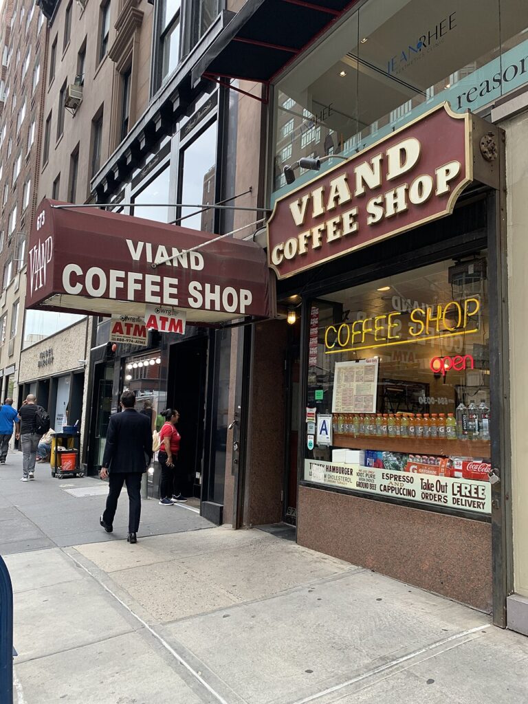 A New York street on which the Viand Coffee Shop sits. It has a brown awning sticking out over the entrance and a brown sign above the window, both with the coffee shop name on. In the window is a yellow neon sign reading “coffee shop” and a red one reading “open.”