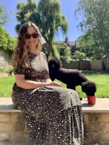 Vicki, a woman with light skin tone and long wavy brown hair, wearing a long leopard print dress and brown sunglasses. She is sat outside on a bright sunny day on a low wall with a lawn behind her on a sunny day, beside her on the wall a small black dog is stood drinking from a red mug.