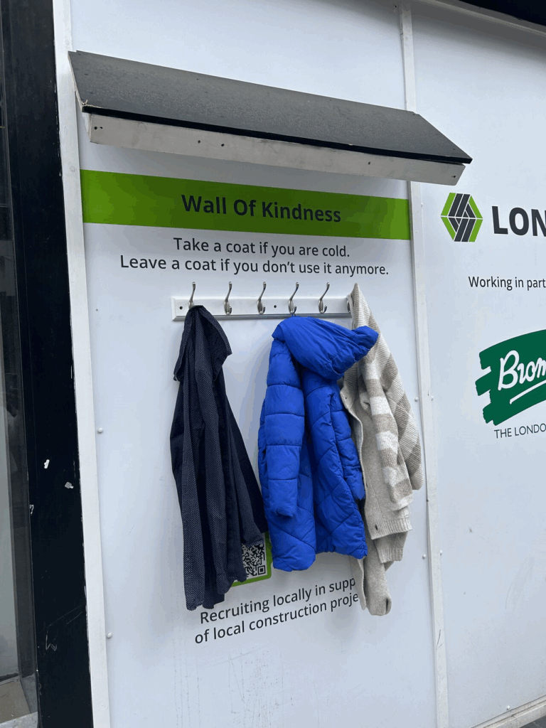 a white wall with a little roof section providing protection over a set of hooks on the wall. There are 3 coats on the set of hooks. Above them text on the wall reads “wall of kindness. Take a coat if you are cold. Leave a coat if you don’t use it anymore.”