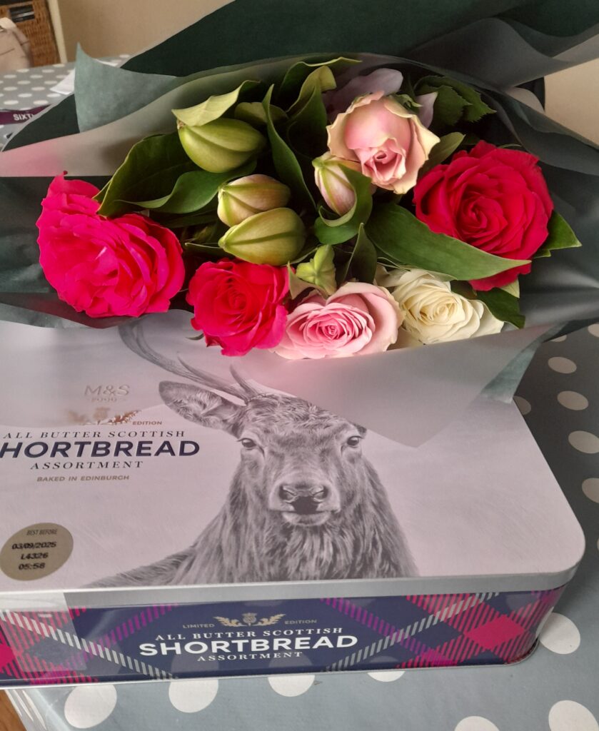 a beautiful bouquet of roses resting on a tin of all butter Scottish shortbread biscuits. On the tin is a black and white illustration of a deer.