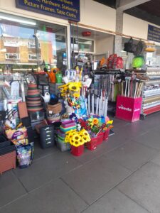 A shop off a street with piles of items outside the door including plant pots, fake sunflowers, brushes, watering cans, and many other items. The part of sign above the shop which is visible reads “hardware, stationery, picture frames, table clothes.”