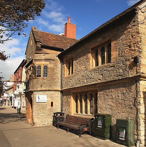 An old building in Bridport of brown stone with wooden windows set in to it. The entrance to it is a rounded turret style which protrudes from the rest of the building. A bend and bin are under the downstairs window. Behind it a pedestrianised street. The sky above is bright blue with a few fluffy clouds.