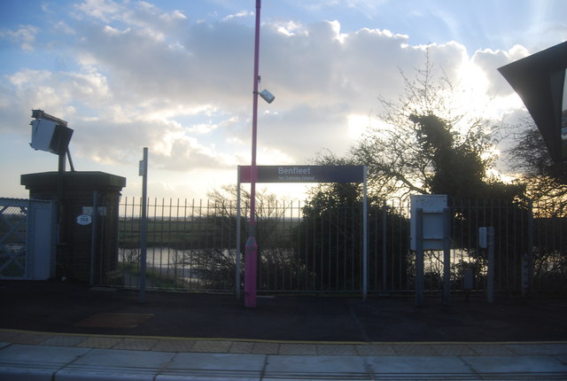 A sign for Benfleet station on the platform at sunset. The shy above has white clouds with some blue sky above it and sun breaking through it as it sets. The platform itself is quite dark.
