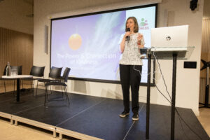 Sarah, a woman with light skin tone and shoulder length light brown hair, standing on a stage holding a microphone. To one side of her is a laptop on a stand. To the other are several empty chairs around a small table. Behind her is a screen showing a slide with text on it reading “the power and connection of kindness.”
