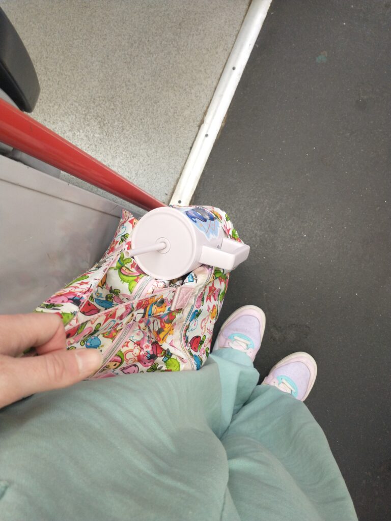 a view down someone standing on a train. They are wearing pale blue dungarees, pastal blue purple and yellow trainers, and holding a brightly multi-coloured bag with a large pale pink reusable water bottle in it.