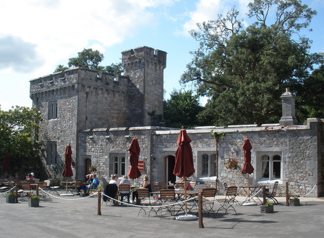 the tea rooms at Powderham Castle. Part of the grey stone castle, most of it just one room tall but a turret to the side has a second floor with a smaller turret attached a little taller. Outside of it are tables and chairs with red parasols folded down. The sky is blue and cloudy