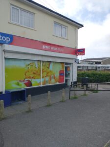 a One Stop shop with the windows covered with a painting of apples and other foods, a red sign along the top reading “great value locally.” The shop is on the corner with the door open.