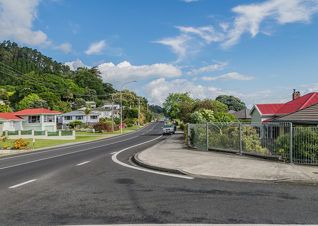 a road in a suburban area of Waikato, New Zealand on a sunny day with wispy white clouds in the sky. On the left side are white buildings with perfectly manicured lawns and a woodland or forest behind them. On the right is a metal fence with bushes the other side of it and more white buildings.
