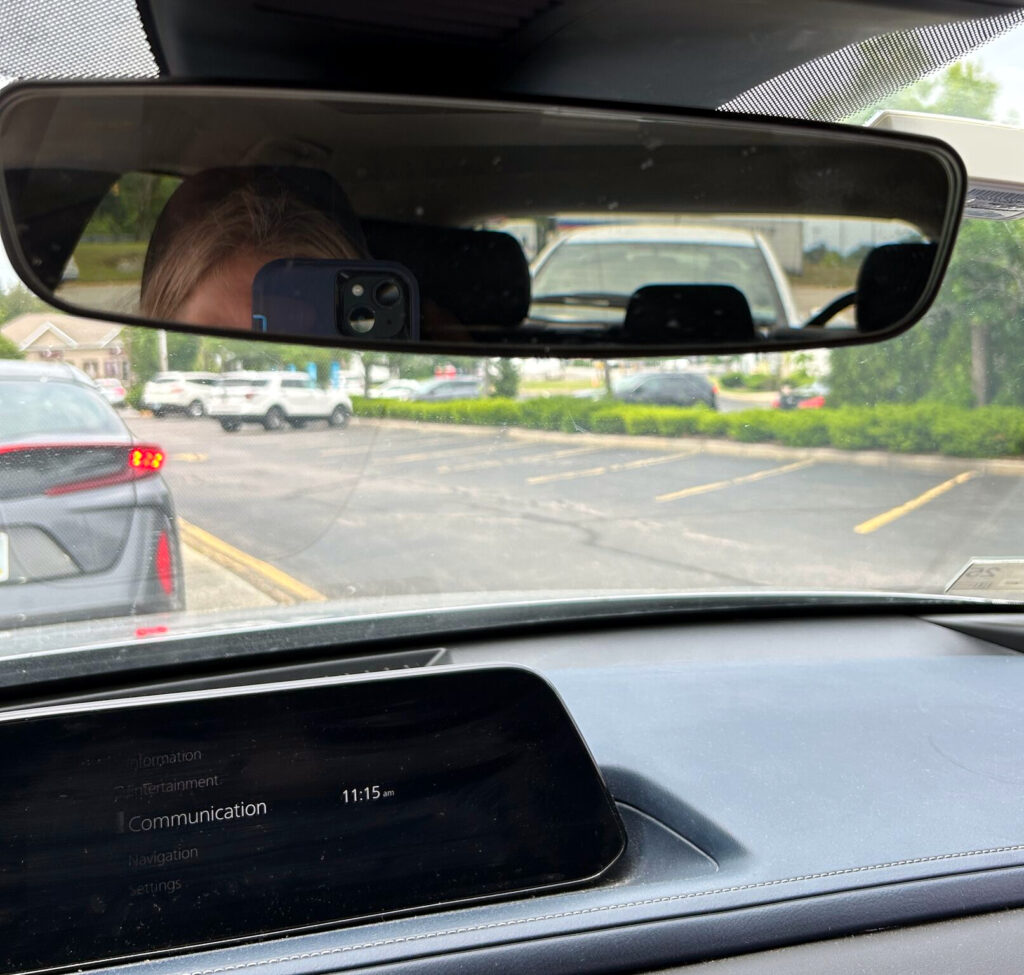 a person with blonde hair and light skin tone taking a photo in the rear view mirror of their car. A white car is visible in the mirror, a black car through the front window. They are in the queue at Wendy's drive thru.