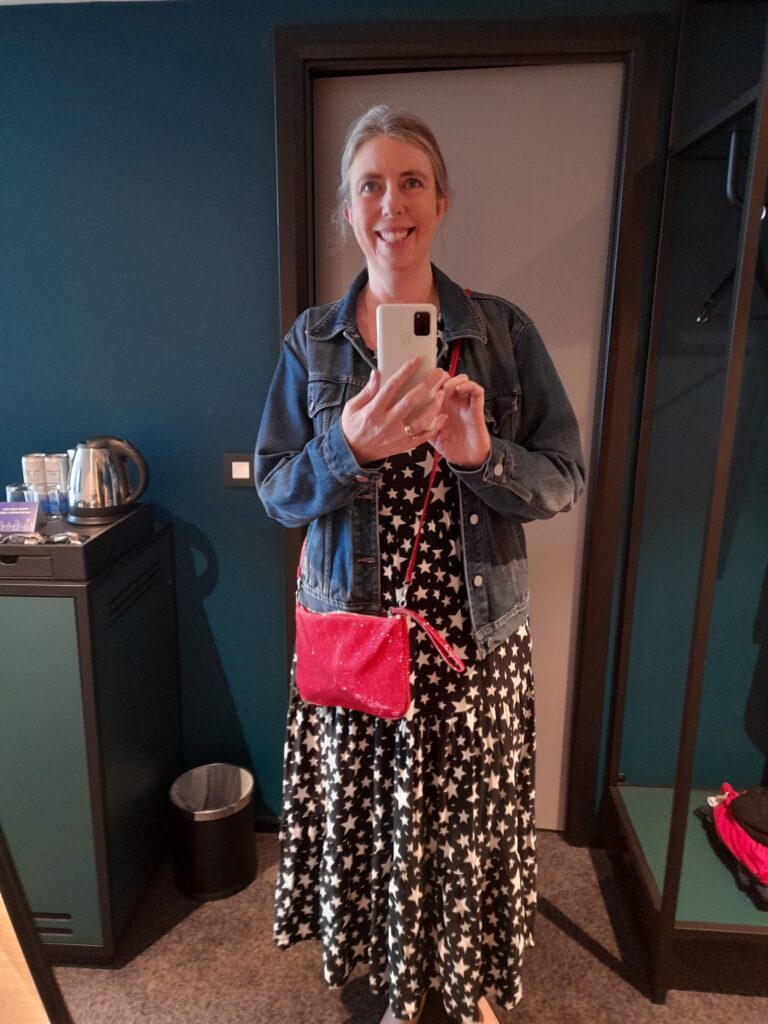 Sarah, a woman with light skin tone and long grey hair tied back wearing a long black dress covered in white stars and a denim jacket with a small pink bag. She is standing in front of a mirror taking a selfie with her phone.