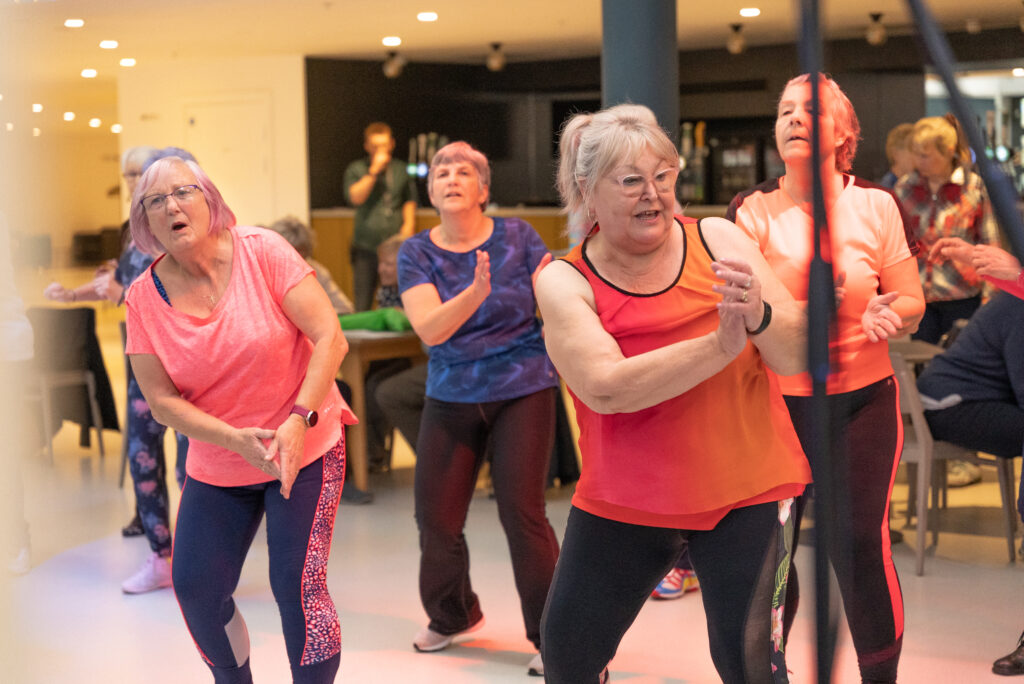 5 older people with light skin tone at an aerobics fitness class. They are wearing fitness gear in bright colours, all leaning to one side and reaching their hands to the other side.