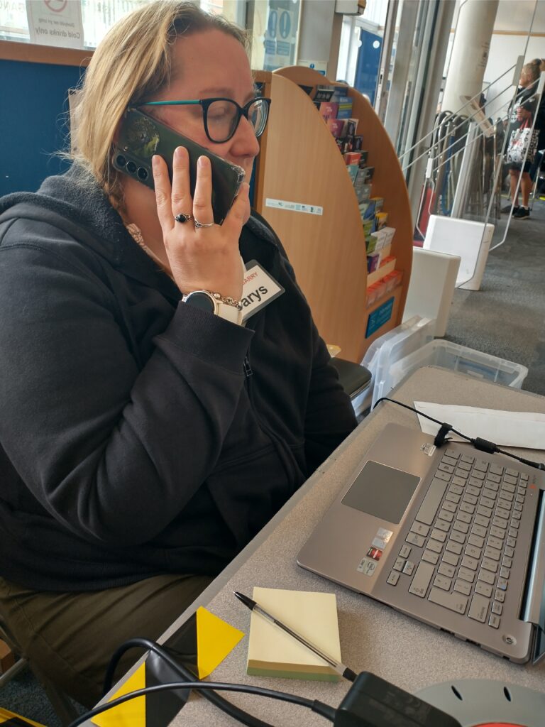 a woman with light skin tone and long blonde hair holding a phone to her ear and speaking into it. She is sat at a table with a laptop in front of her in a library.
