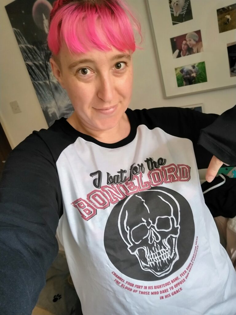 a woman with light skin tone and bright pink hair tied back with some in a fringe on her face smiling and pointing to the design on her top. The top has long sleeves which are black and a white body with “I bat for the bonelord” on it and a large black circle with a skull on it.
