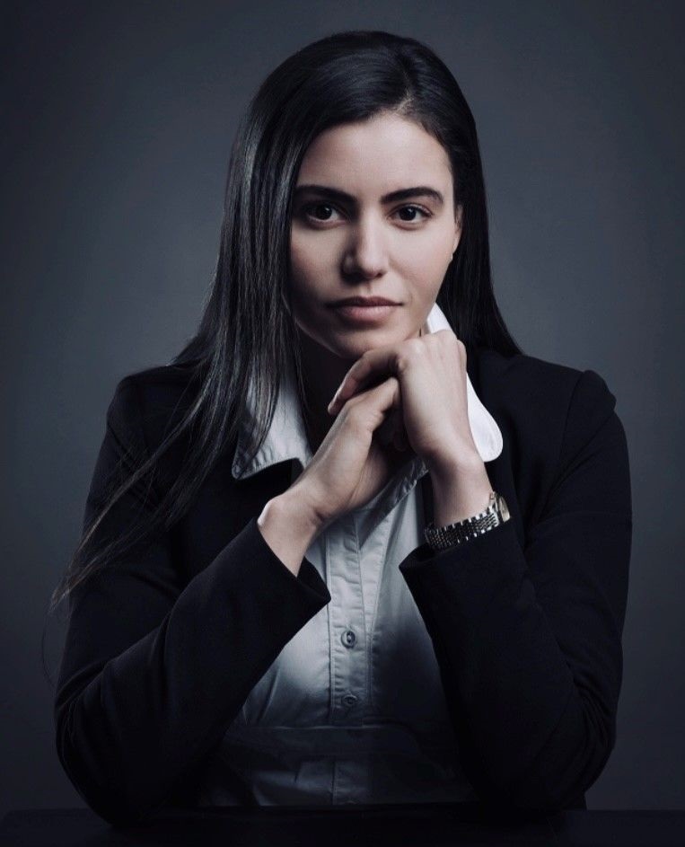 Sara, a woman with light skin tone and long dark hair, wearing a white shirt and a black blazer. She is sat down with her elbows on a table and hands resting together just in front of her face.
