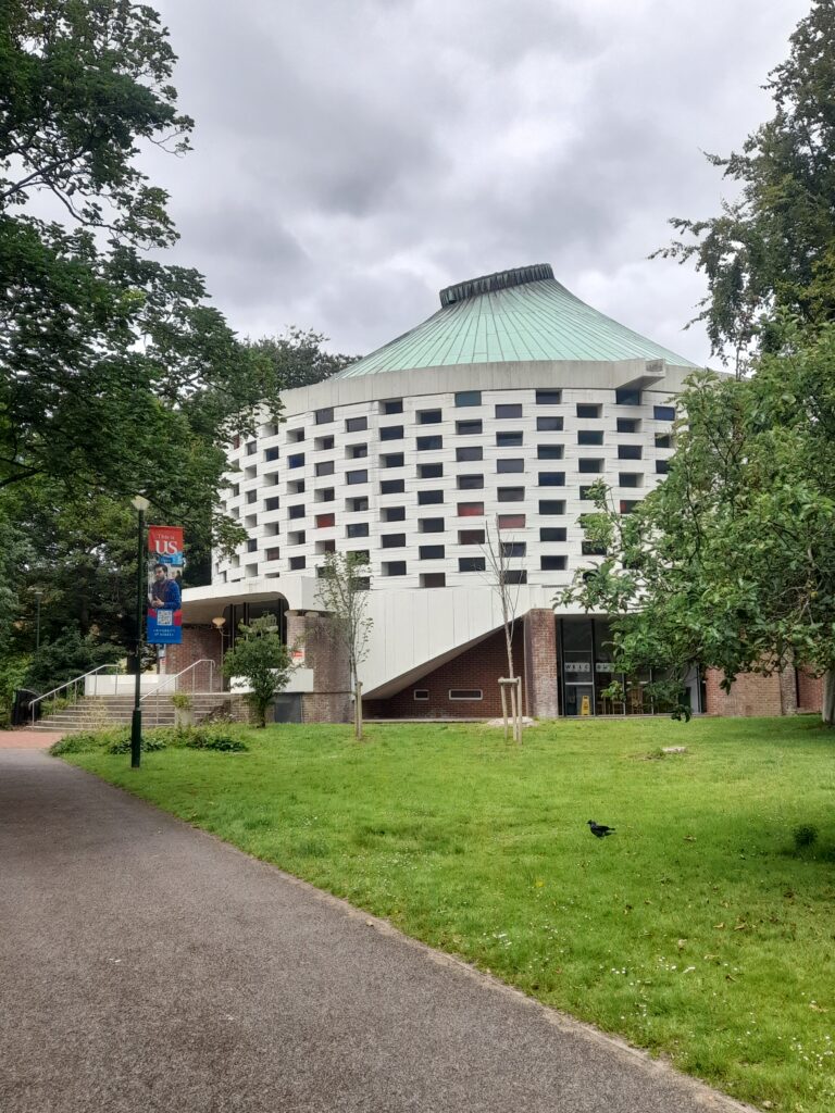 A building at the Universiry of Sussex. It is round with lots of windows and a tipi style green roof. Around it is a grassy area and trees.