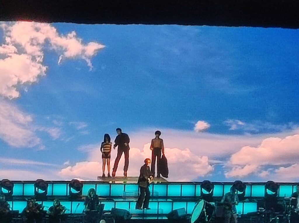 4 members of Pulp on stage at Glasgow OVO hydro arena with a screen showing blue sky with fluffy clouds behind them. 3 of the people are standing at the top of the stage while one person walks down some steps going across the stage which are lit up blue.
