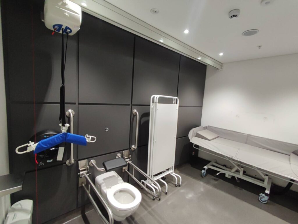 A large disabled toilet with a changing space, the red pull cord all the way to the floor, and a hoist.