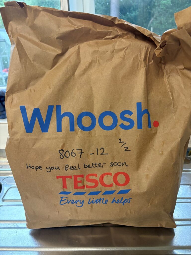 A large brown paper bag with “Tesco Whoosh” printed on it. In handwritten text is an order number and “hope you feel better soon.”