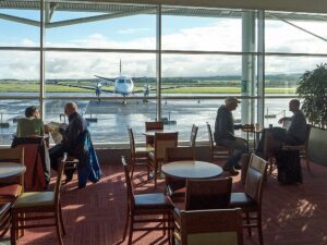 A restaurant at Inverness airport looking out onto the tarmac through a huge window where a plane is parked. People are sat at a couple of tables eating, drinking and reading a newspaper.