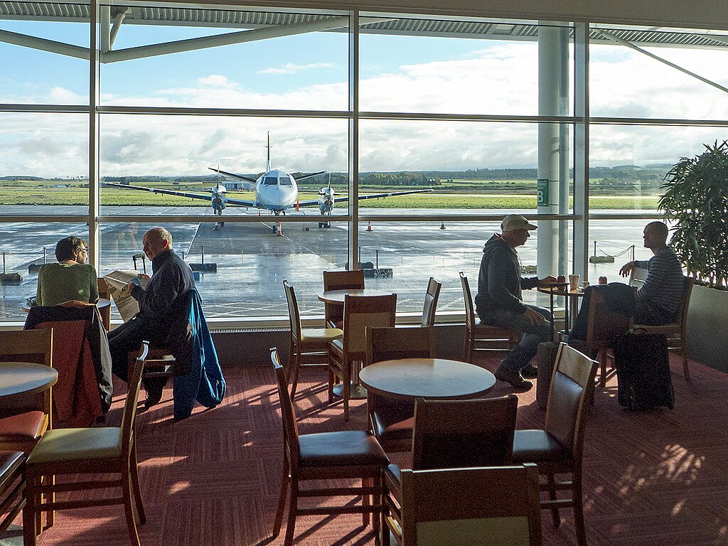 A restaurant at Inverness airport looking out onto the tarmac through a huge window where a plane is parked. People are sat at a couple of tables eating, drinking and reading a newspaper.
