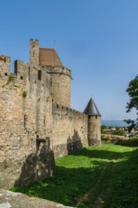 To the left are very old walls around a castle or city. A small round turret at the corner. To the right is a grassy area shaded by trees. The sky is bright blue and cloudless and past the grass miles of greenery and forests are visible out to the horizon.